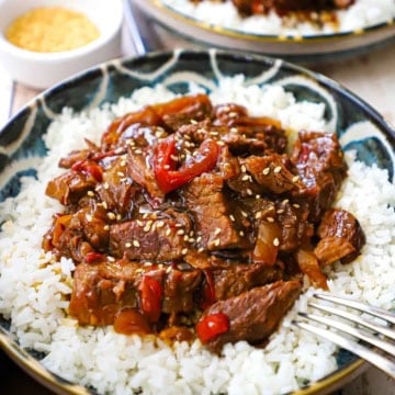 A close-up view of slow cooker pepper steak on top of steamed white rice on colorful shallow bowl.
