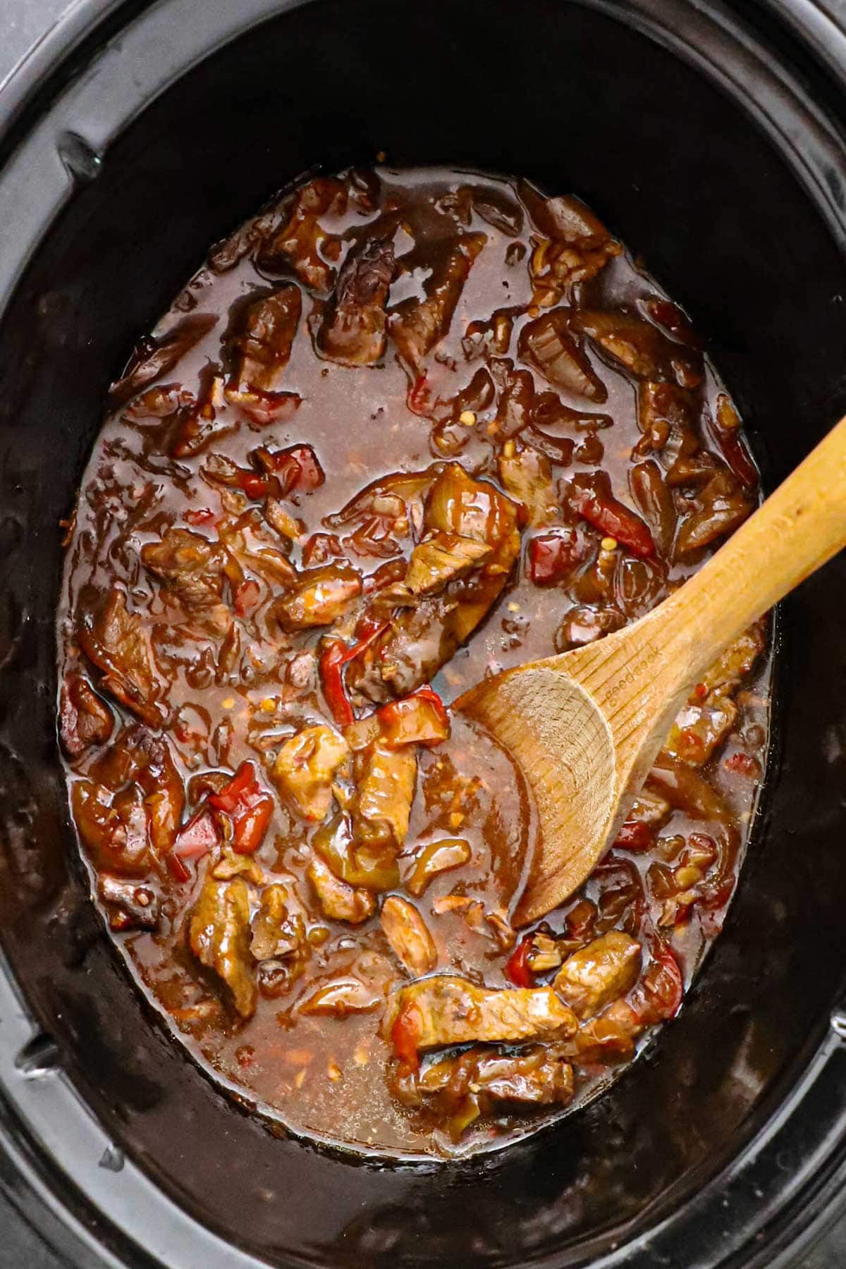An overhead view of the inside of a slow cooker that is filled with pepper steak in a dark brown gravy with a wooden spoon in the middle of the sauce.