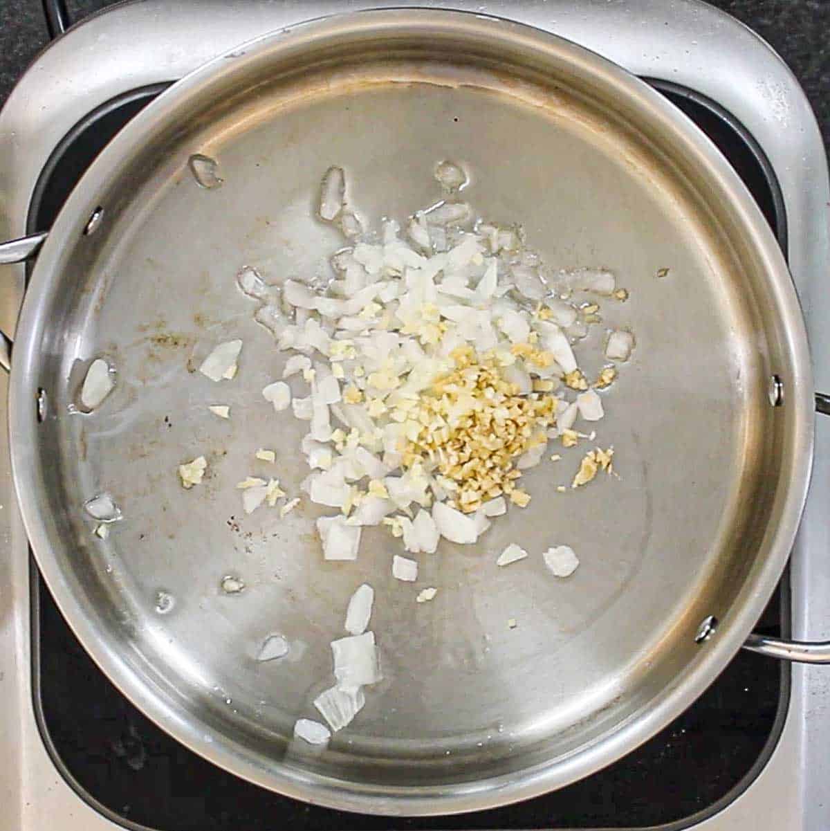 A large silver skillet filled with chopped onion, minced garlic, and minced ginger being sautéed in olive oil.