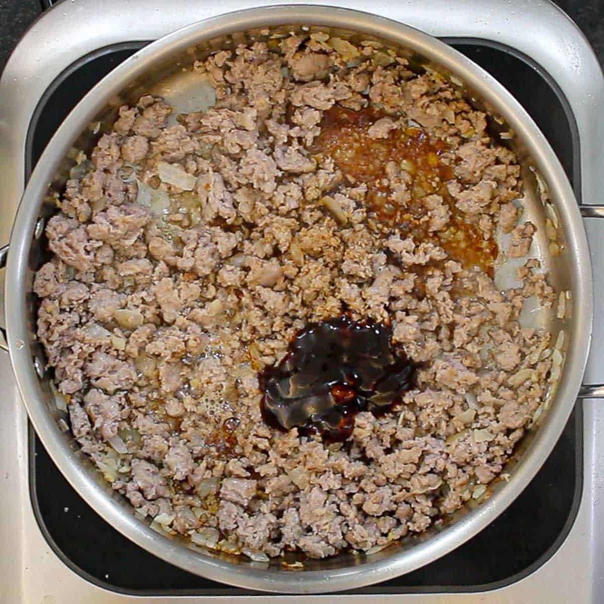 A skillet filled with ground pork that has been fully cooked with sautéed onion, garlic, and ginger with a pool of hoisin sauce resting on top of the meat mixture.