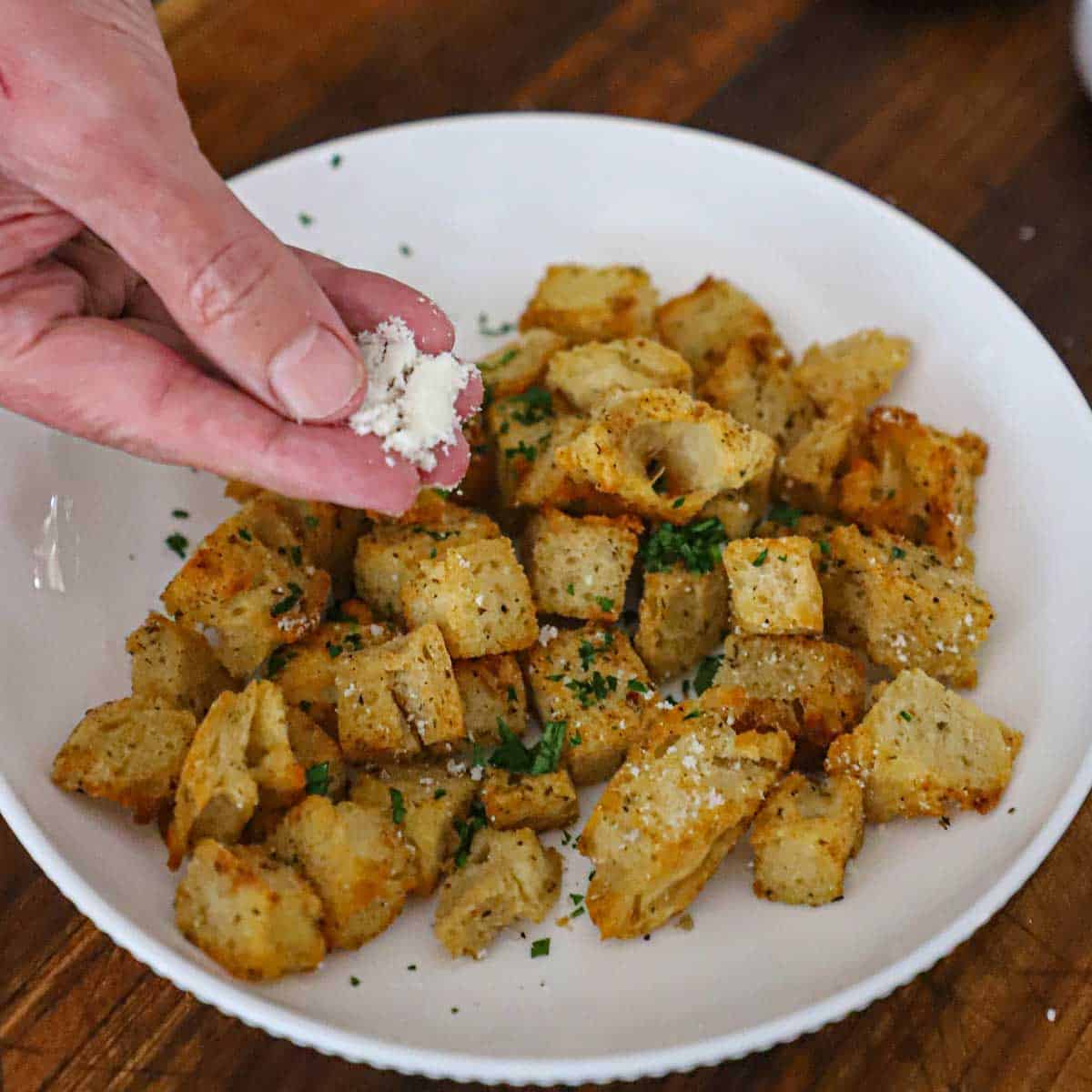 A person sprinkling grated Parmesan over the tops of freshly baked homemade croutons in a white bowl.