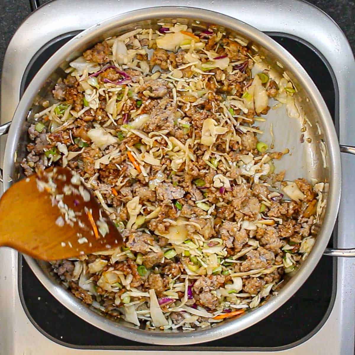 A person using a wooden spatula to stir a pork and cabbage mixture in a skillet that will be used as the filling for homemade egg rolls.