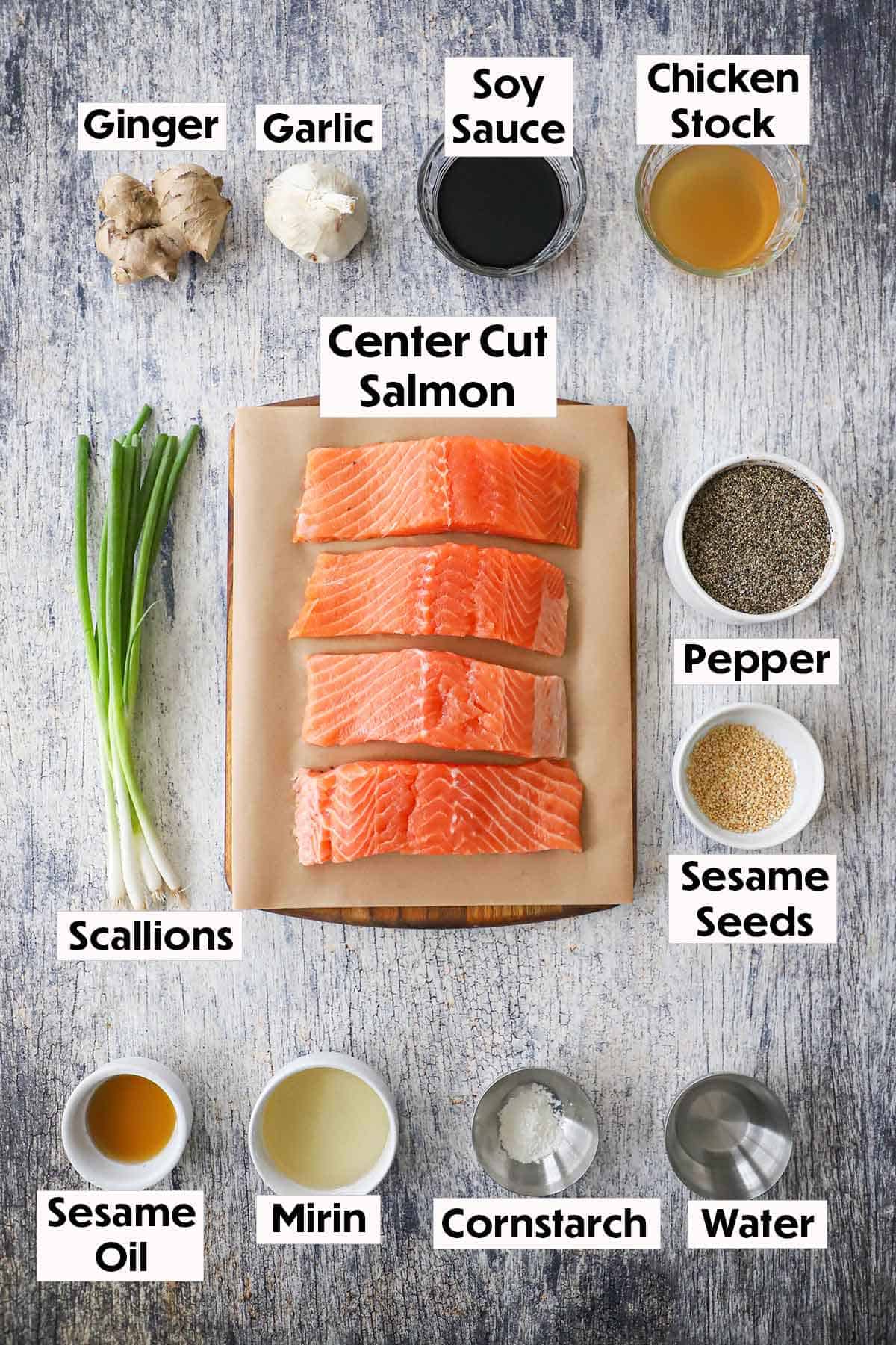 An arrangement of ingredients on a grey wooden background including center-cut salmon fillets, ginger, garlic, soy sauce, chicken stock, scallions, and seasonings.