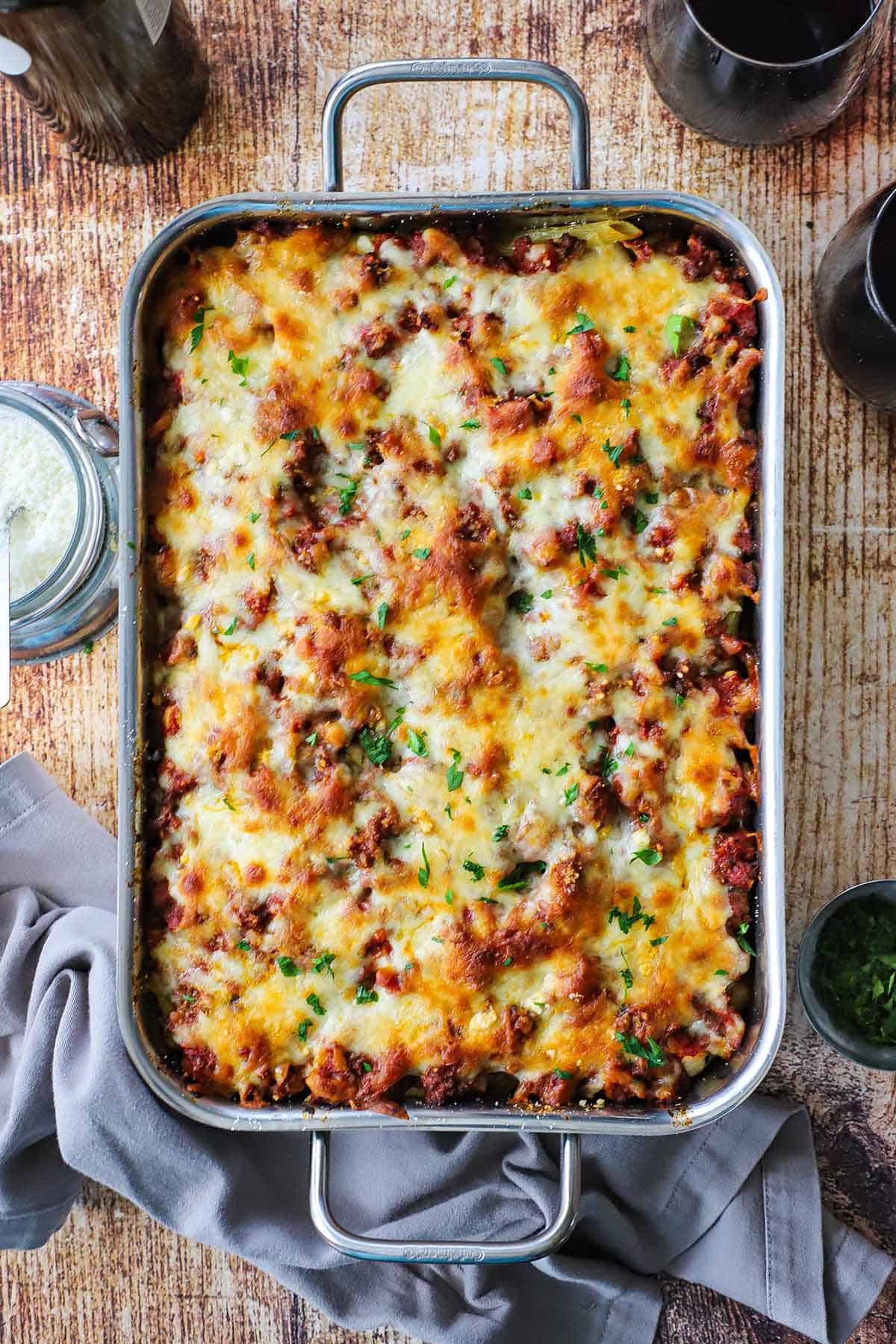 A large stainless steel lasagna pan filled with a freshly baked best-ever baked ziti surrounded by a couple glasses of red wine and a bottle of wine.