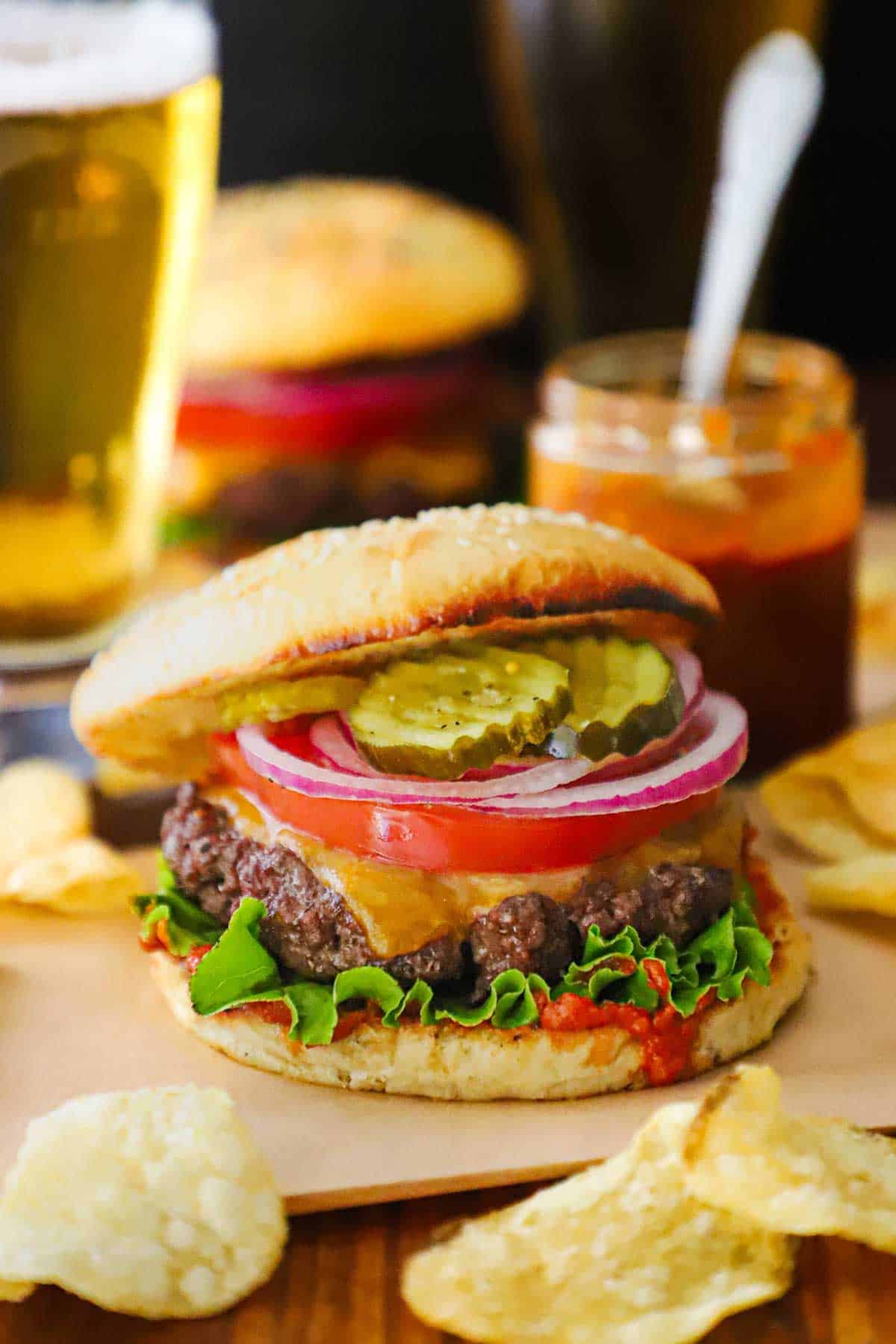 A best-ever cheeseburger topped with a slice of tomato, thin slices of red onion, bread and butter pickles, resting on green leaf lettuce, homemade ketchup, all inside a homemade hamburger bun.