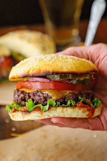 A person holding a best-ever cheeseburger consisting of a juicy beef burger topped with melting cheddar cheese, lettuce, tomato, red onion, and pickles all sandwiched between a toasted homemade hamburger bun.