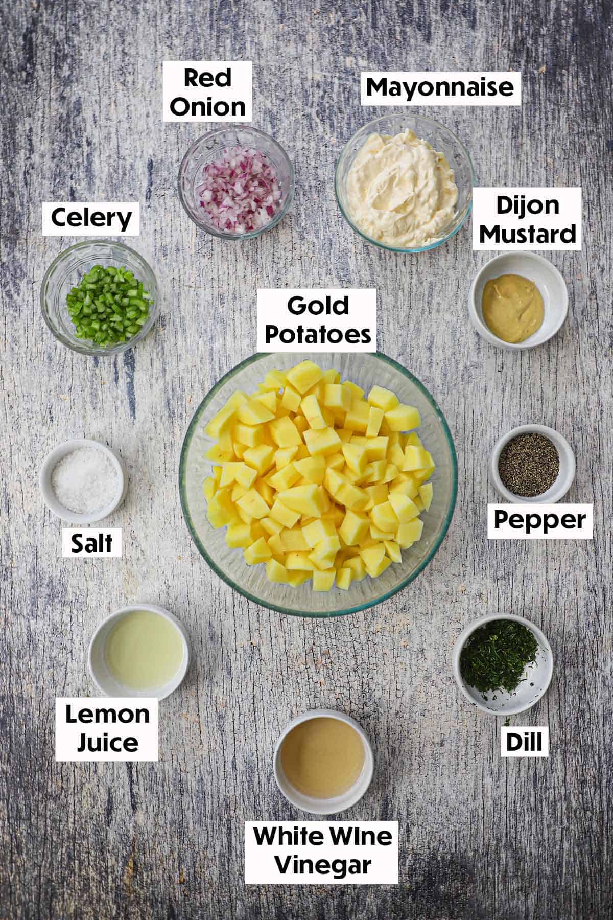 An arrangement of ingredients for best-ever potato salad on a grey wooden background including bowls filled with red onion, celery, mayonnaise, Dijon, cubed potatoes, lemon juice, vinegar, chopped dill, salt, and pepper.