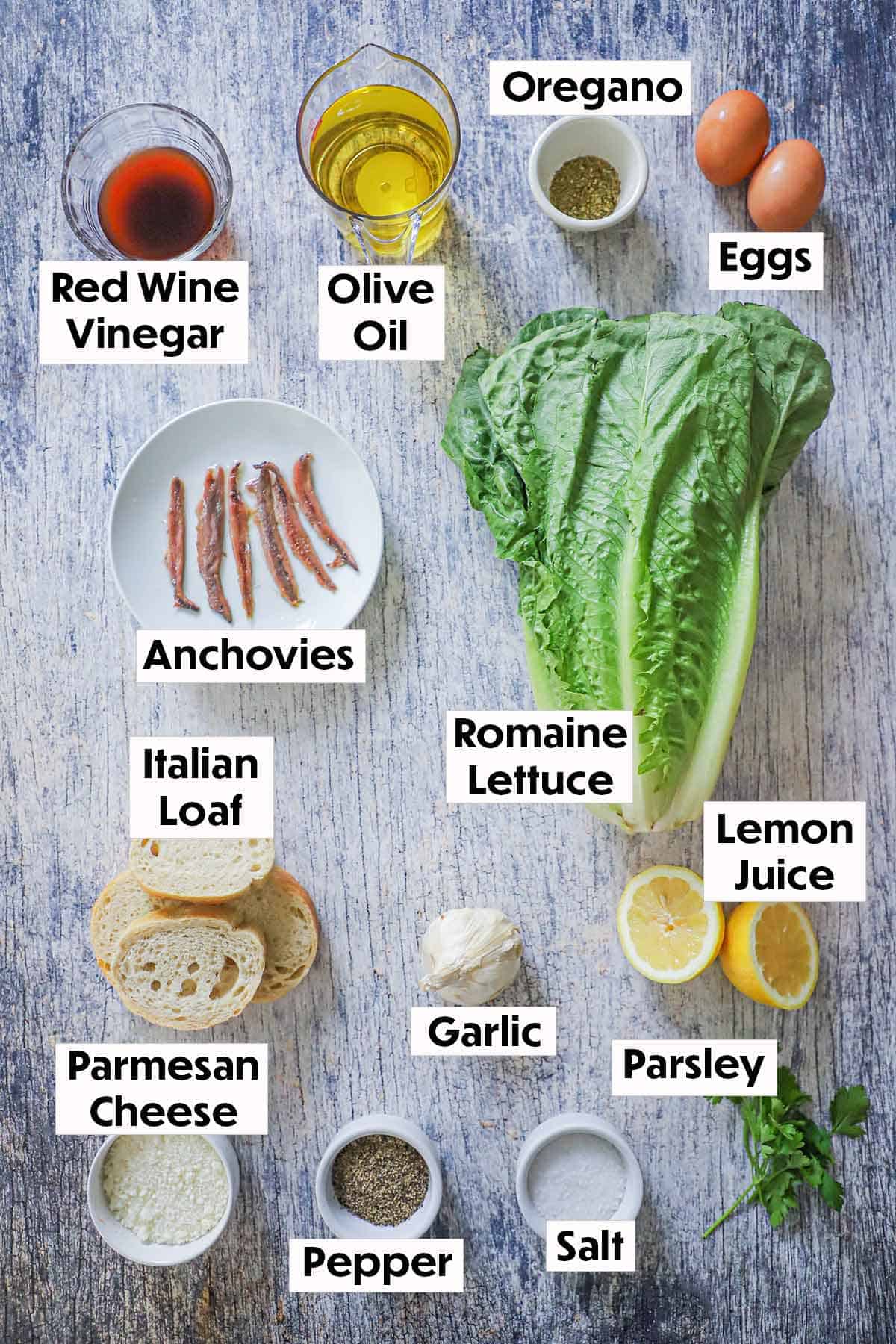 An arrangement of ingredients for classic Caesar salad on a grey wooden background including romaine lettuce, vinegar, oil, eggs, anchovies, herbs, bread, lemon juice, garlic, and fresh parsley.