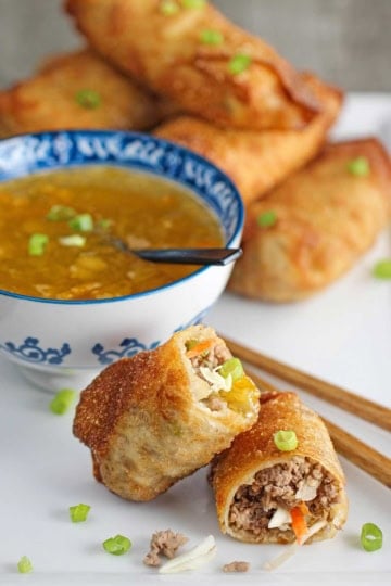 A homemade egg roll that has been split in half revealing the pork and cabbage inside with one of the halves resting on the other half with a bowl of homemade duck sauce in the background.