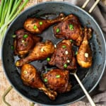 A large non-stick skillet filled with Filipino adobo chicken with a thick sauce over the chicken and in the bottom of the pan and topped with chopped scallions.