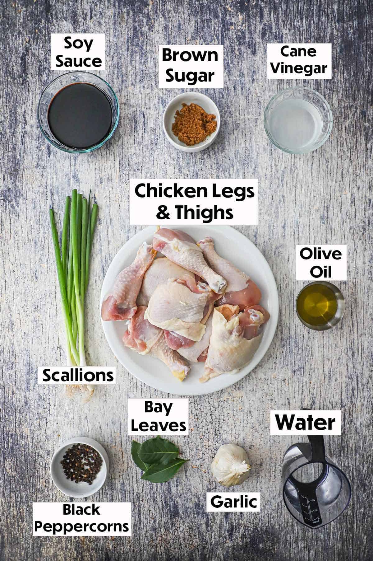 An arrangement of ingredients for Filipino chicken adobo on a grey wooden background including raw chicken thighs and legs, soy sauce, brown sugar, vinegar, scallions, oil, peppercorns, bay leaves, and garlic.