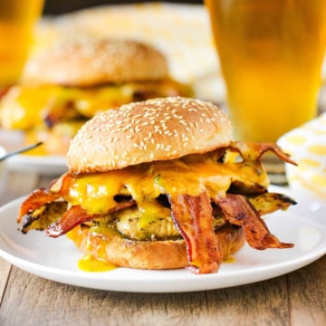 A grilled honey mustard chicken sandwich with bacon on a sesame seed bun sitting on a withe dinner plate with a frosty glass of beer in the background.