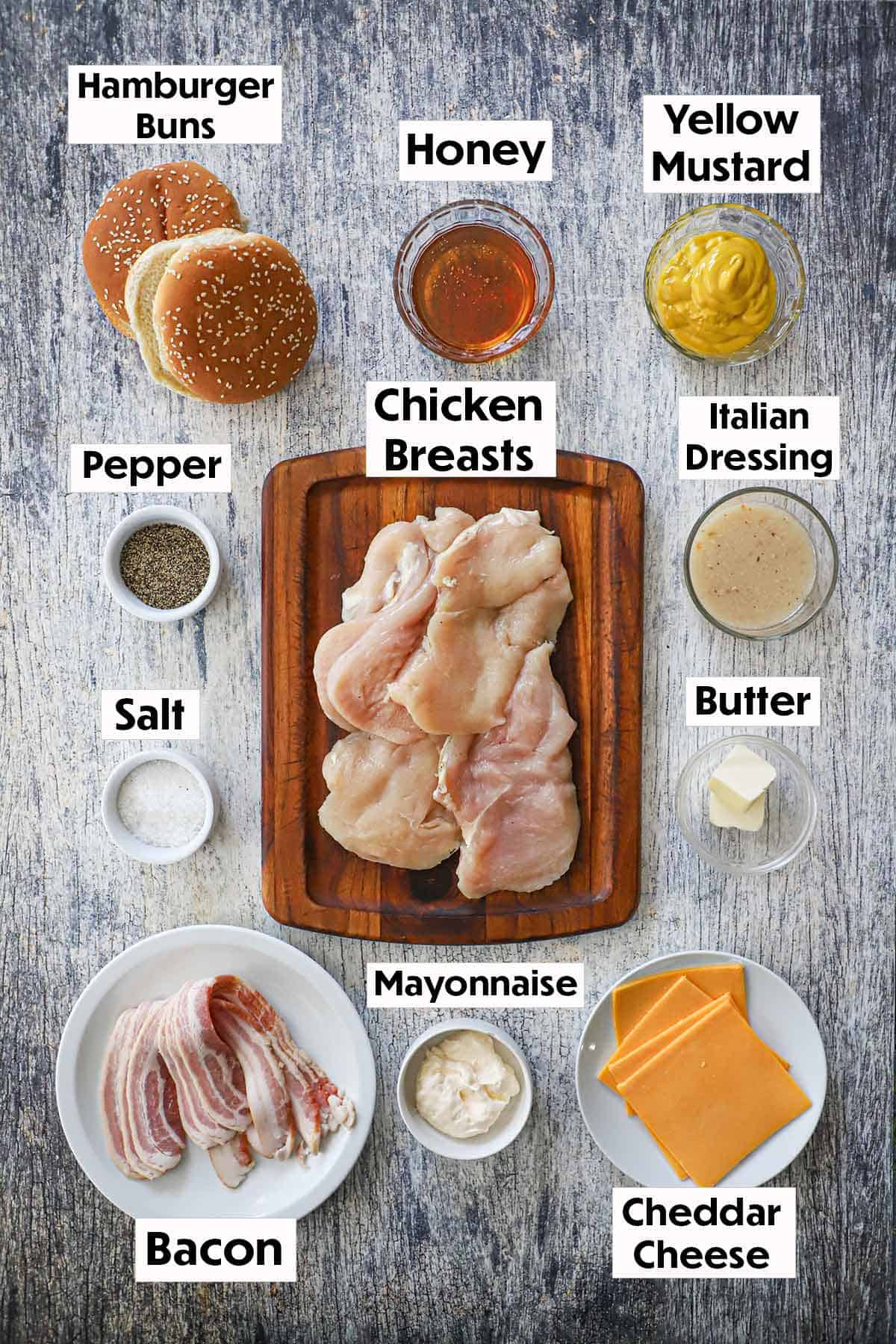 An arrangement of ingredients for a grilled honey mustard chicken sandwich on a wooden grey background including uncooked chicken breasts, hamburger buns, honey, yellow mustard, Italian dressing, seasonings, bacon, mayonnaise, cheddar cheese slices, and butter.