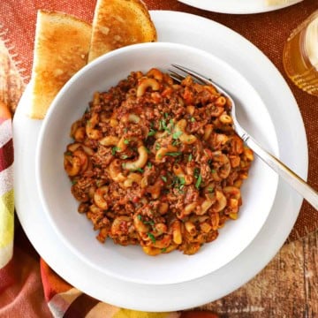 A white bowl filled with Mom's goulash (American-style) sitting on a white dinner plate with two pieces of toast resting on the plate next to the bowl.