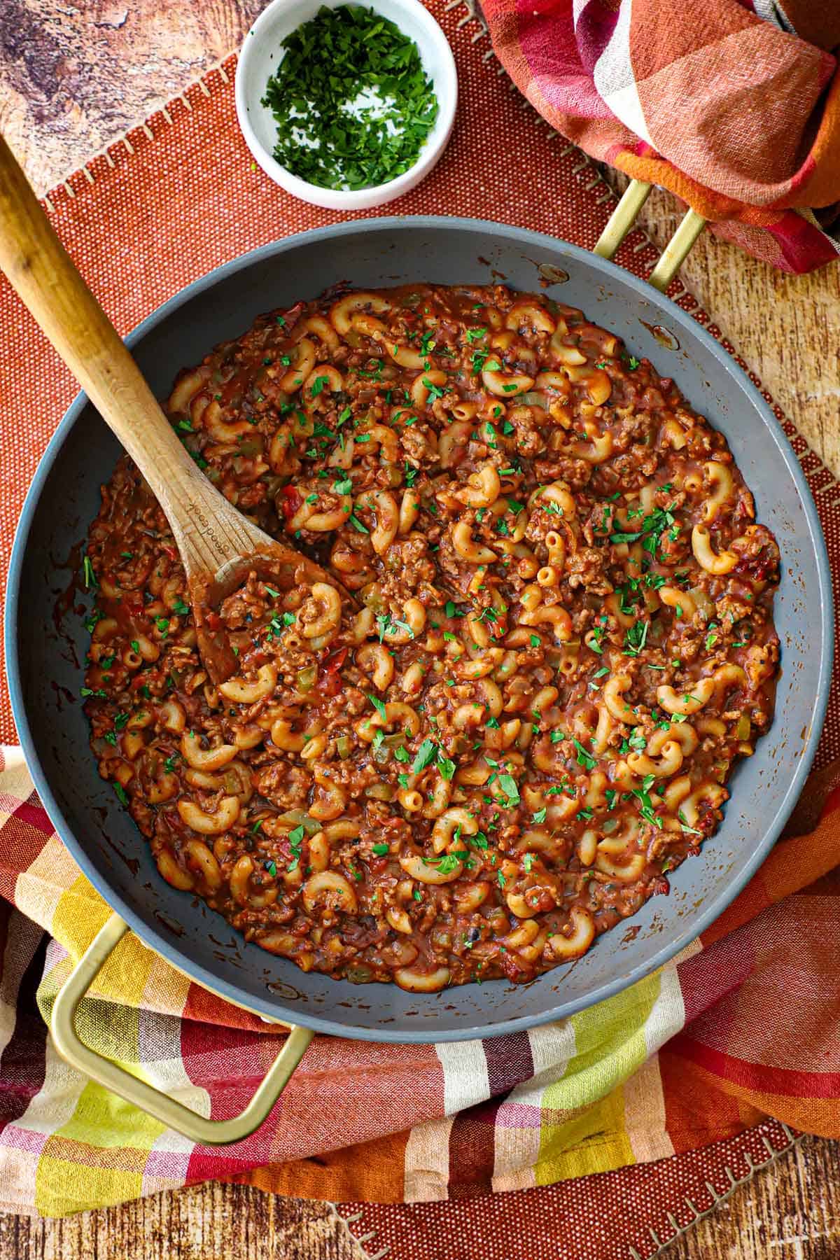 A deep skillet filled with Mom's goulash (American-style) with a wooden spoon inserted into it with a small bowl of chopped parsley sitting nearby.