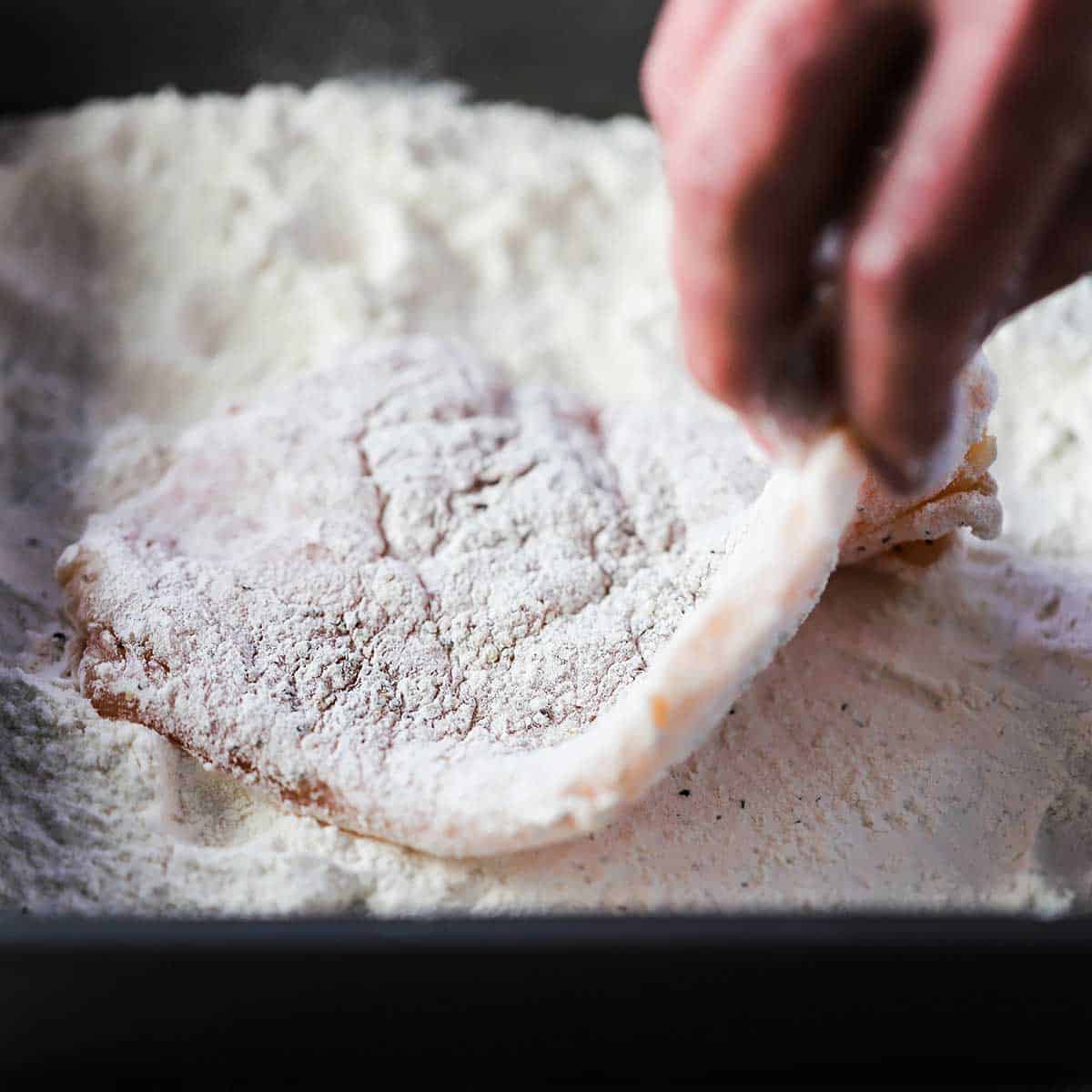 A person holding the edge of a thin uncooked chicken cutlet that has been dredged through a pan full of seasoned flour.