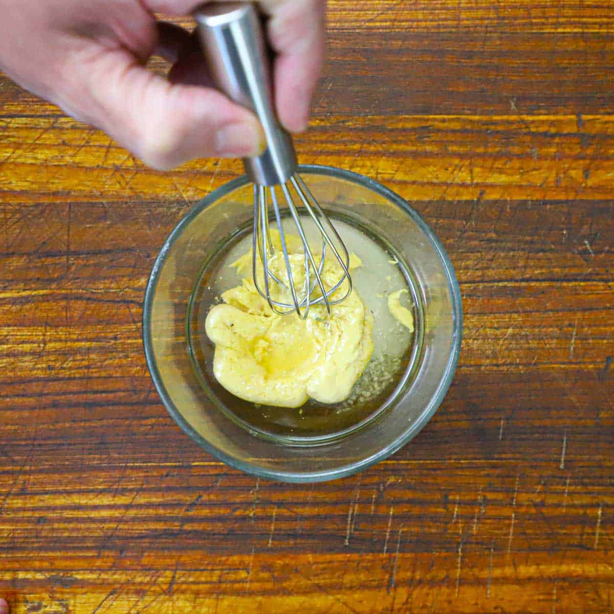 A person using a small whisk to mix vinegar, Dijon mustard, salt, and sugar in a small glass bowl.