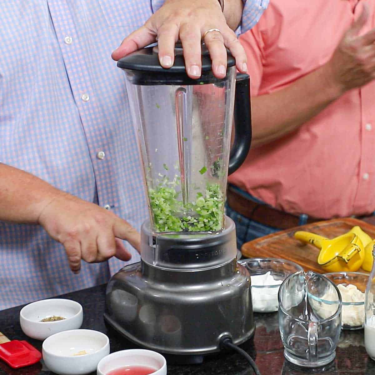 A person using a blender to chop fresh scallions with small bowls of vinegar and spices nearby.