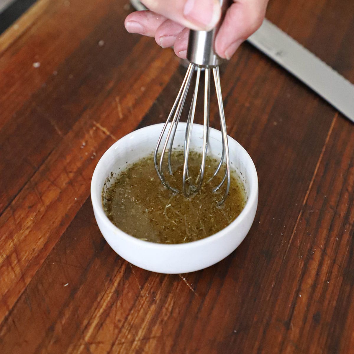 A person using a small whisk to mix oil oil with spices and dried herbs in a small white bowl on a wooden cutting board.