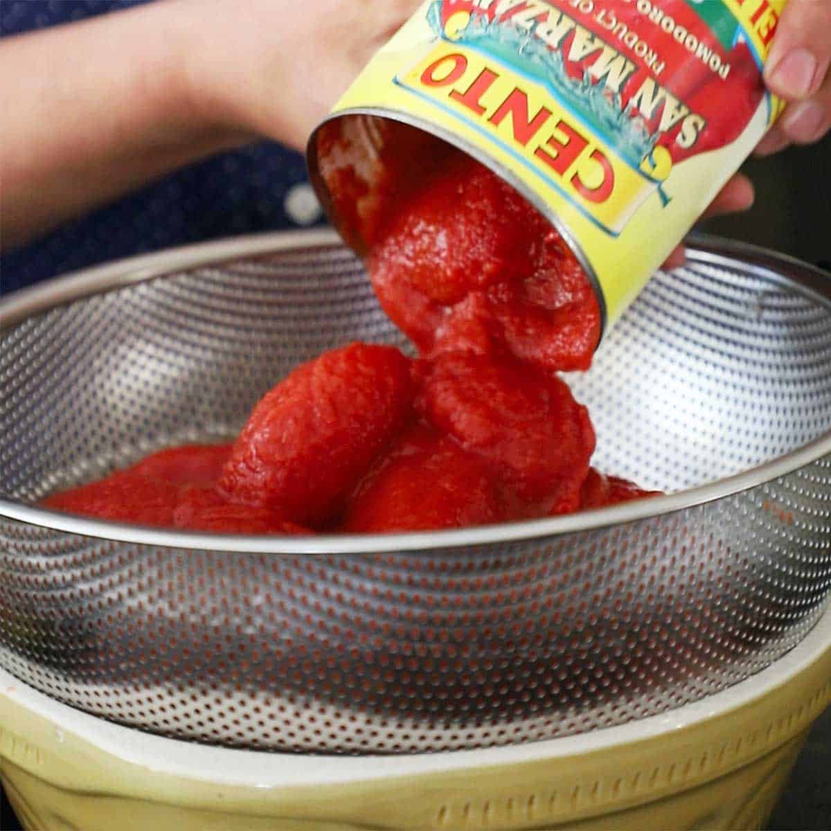 A person dumping whole San Marzano tomatoes from a 28 ounce can into a metal colander resting over a ceramic bowl.