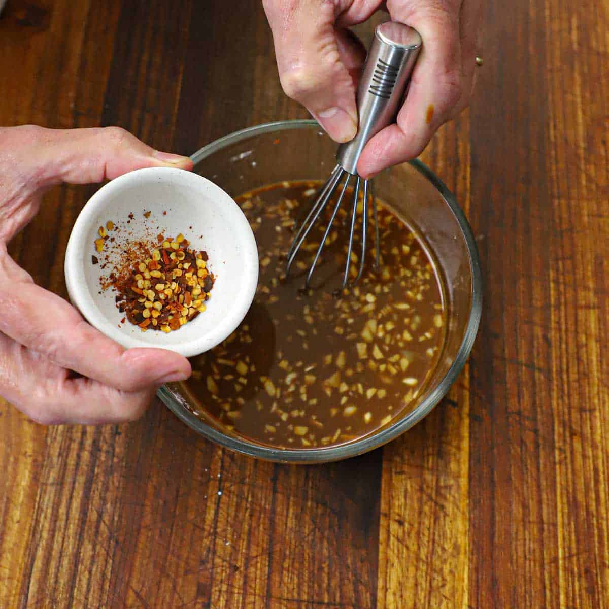 A person using a small whisk to stir a bowl filled with pepper steak sauce while holding a small bowl of red pepper flakes nearby.