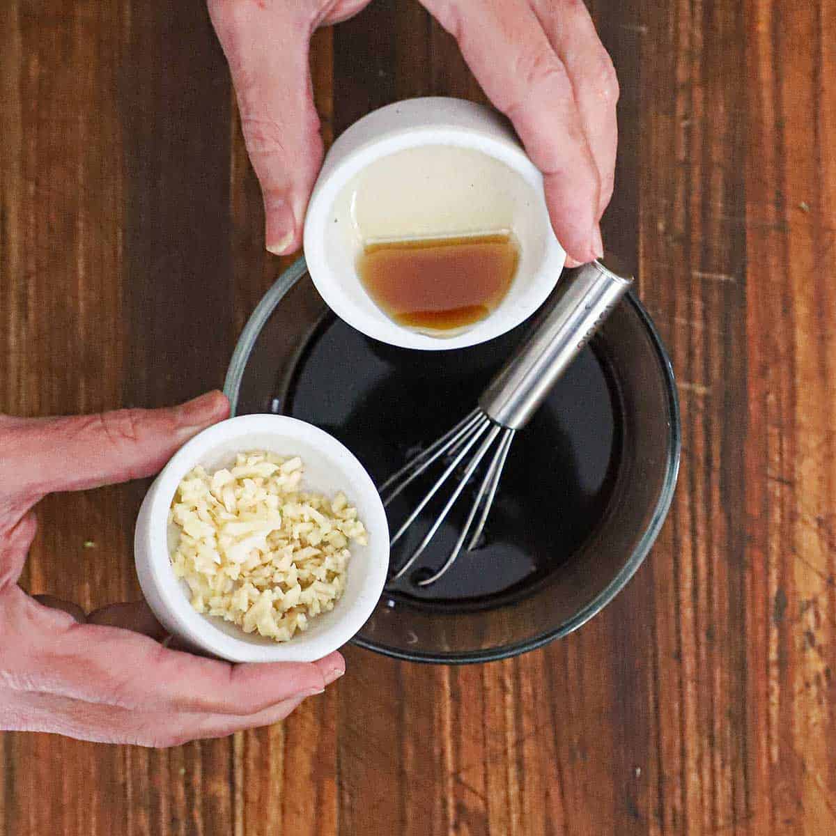 A person holding a small bowl of sesame oil in one hand and another small bowl filled with minced garlic and ginger over a medium-sized bowl filled with a teriyaki sauce.