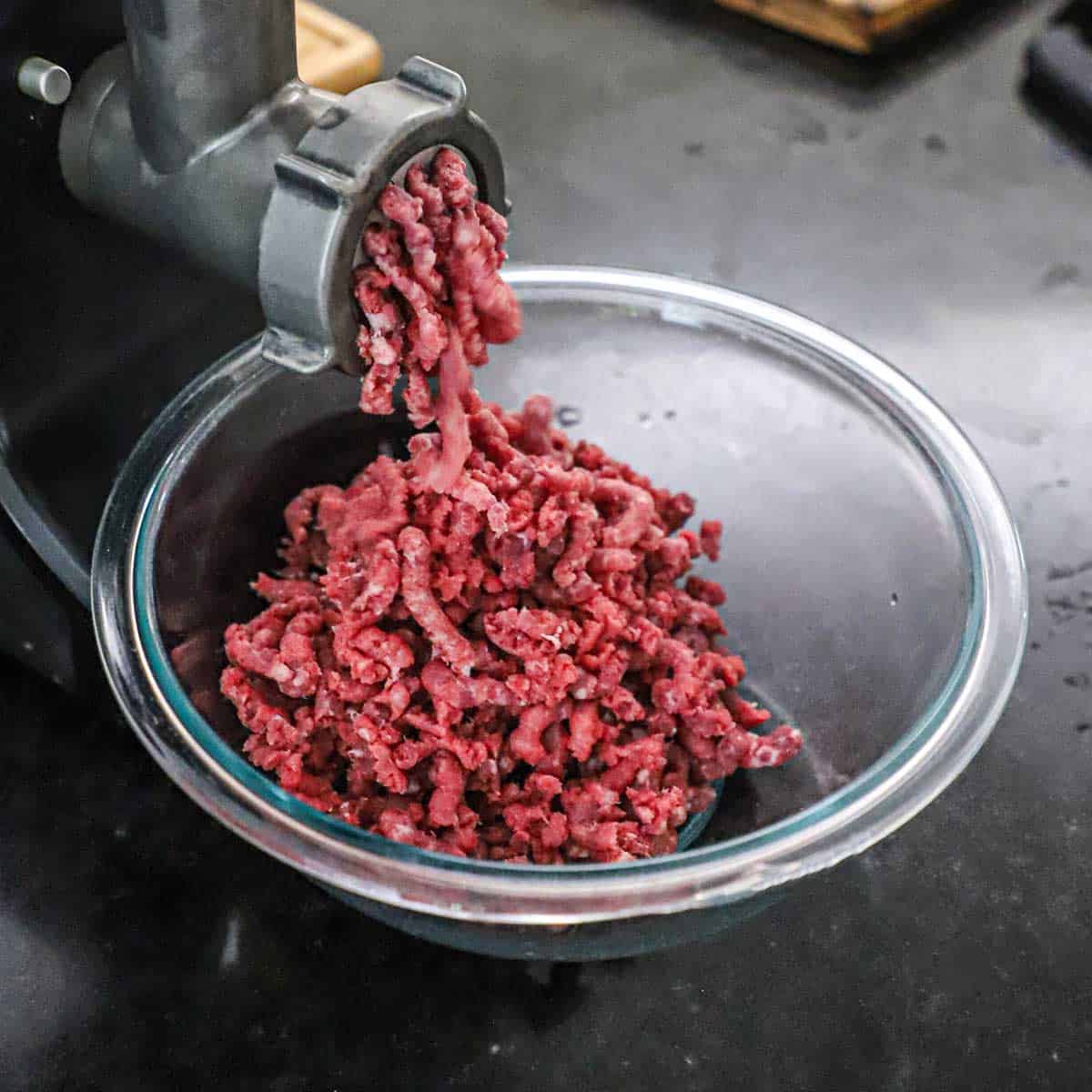 Chunks of skirt steak and flat iron steak being put through a meat grinder with the coarsely ground meat dropping into a glass bowl on a black marble counter.