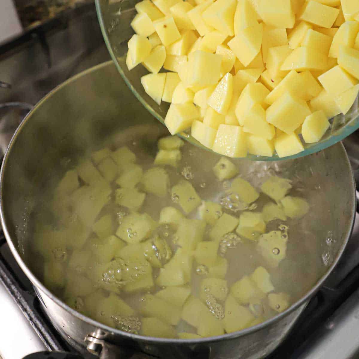 A person dumping peeled and cubed yellow potatoes into a pot filled with boiling water.