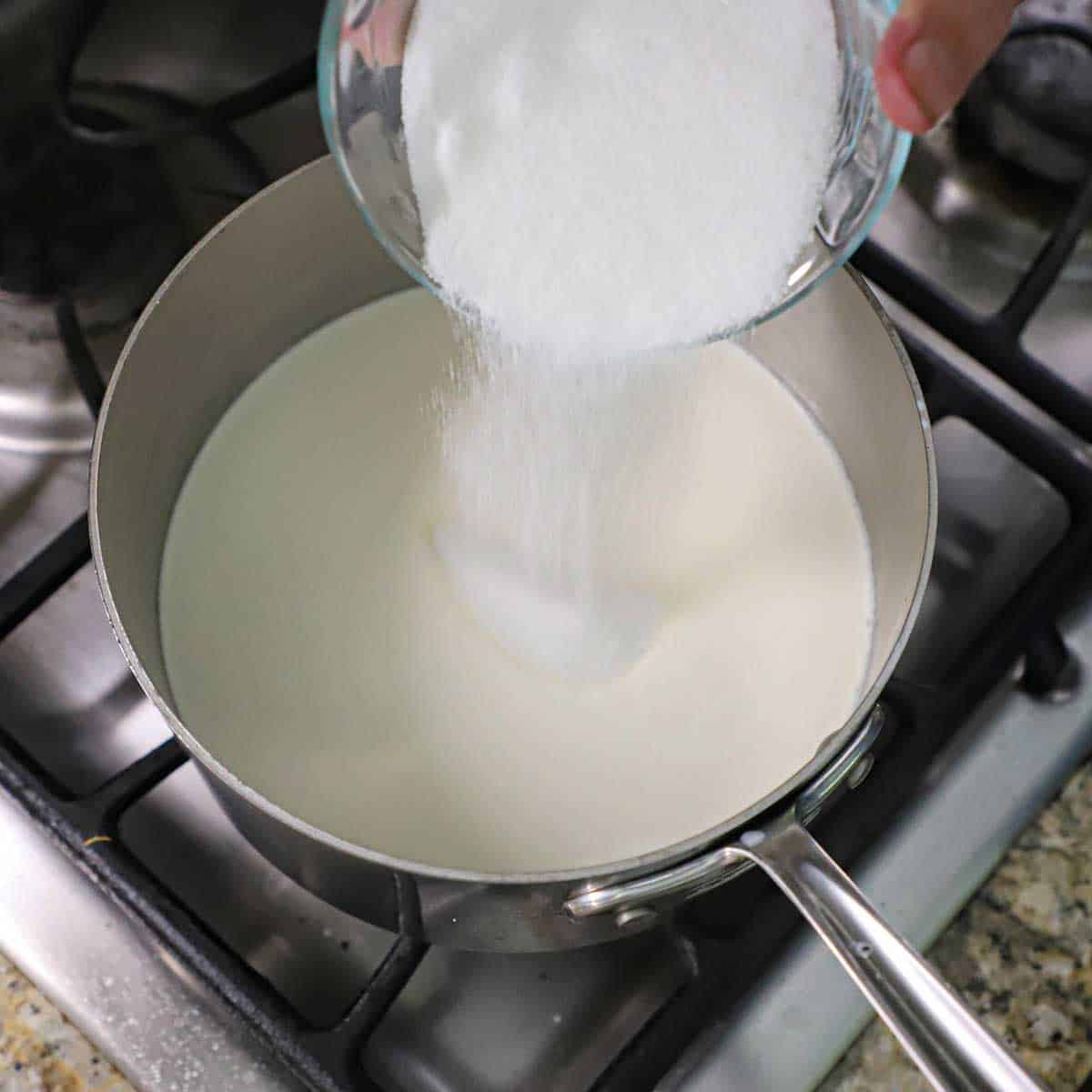 A person transferring granulated sugar into a saucepan filled with simmering cream and milk.