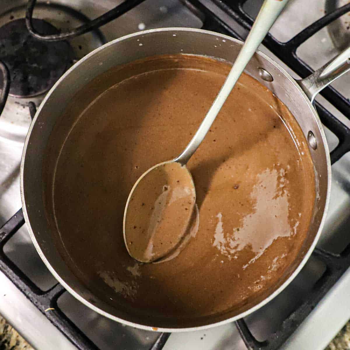 A person using a spoon to lift a mixture of chocolate and simmering milk and heavy cream in a saucepan on a gas stove.