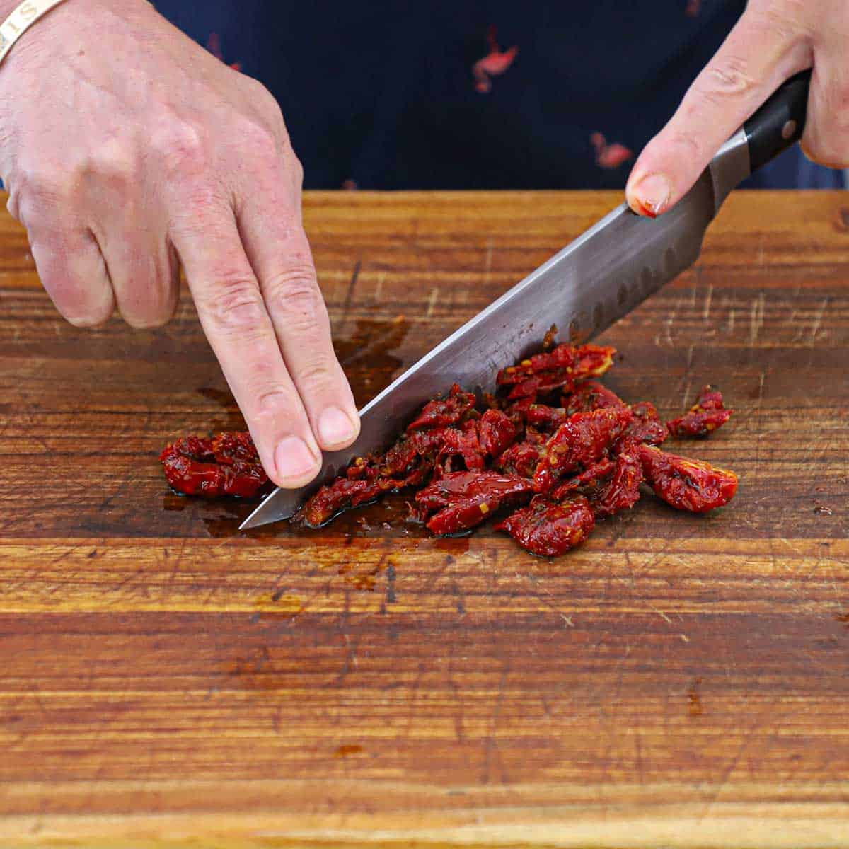 A person using a large chef's knife to roughly chop sun-dried tomatoes on a large wooden cutting board.