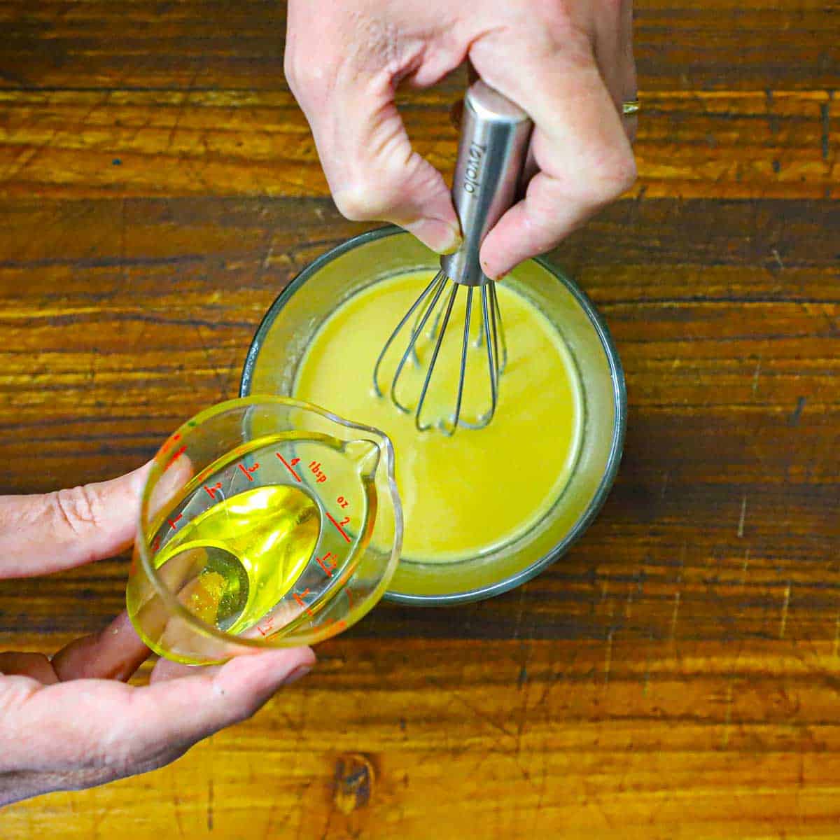 A person using a whisk to emulsify olive oil that is being poured into a bowl of a Dijon vinaigrette.