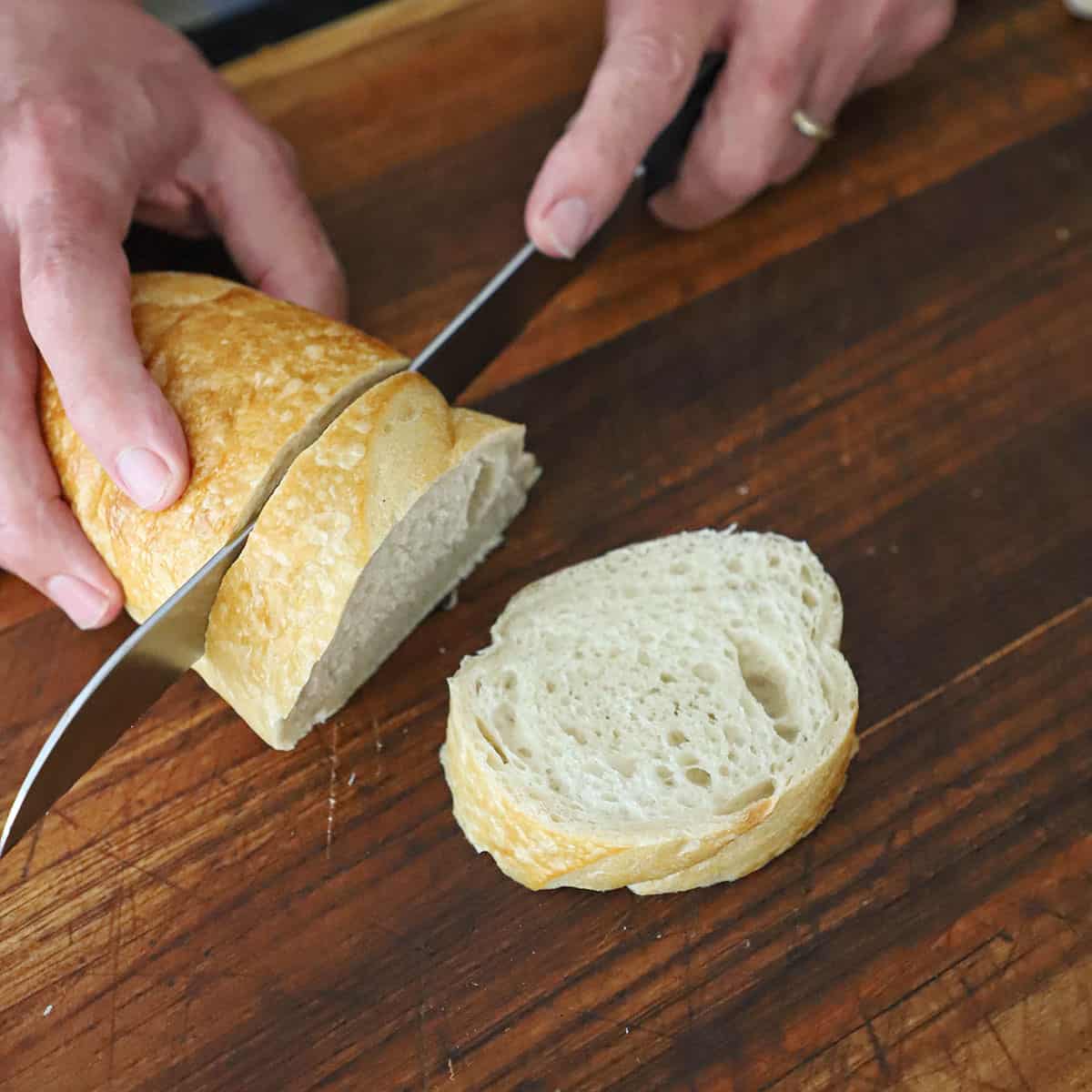 A person using a large serrated knife to slice an Italian loaf of bread into 1-inch thick slices on a wooden cutting board.