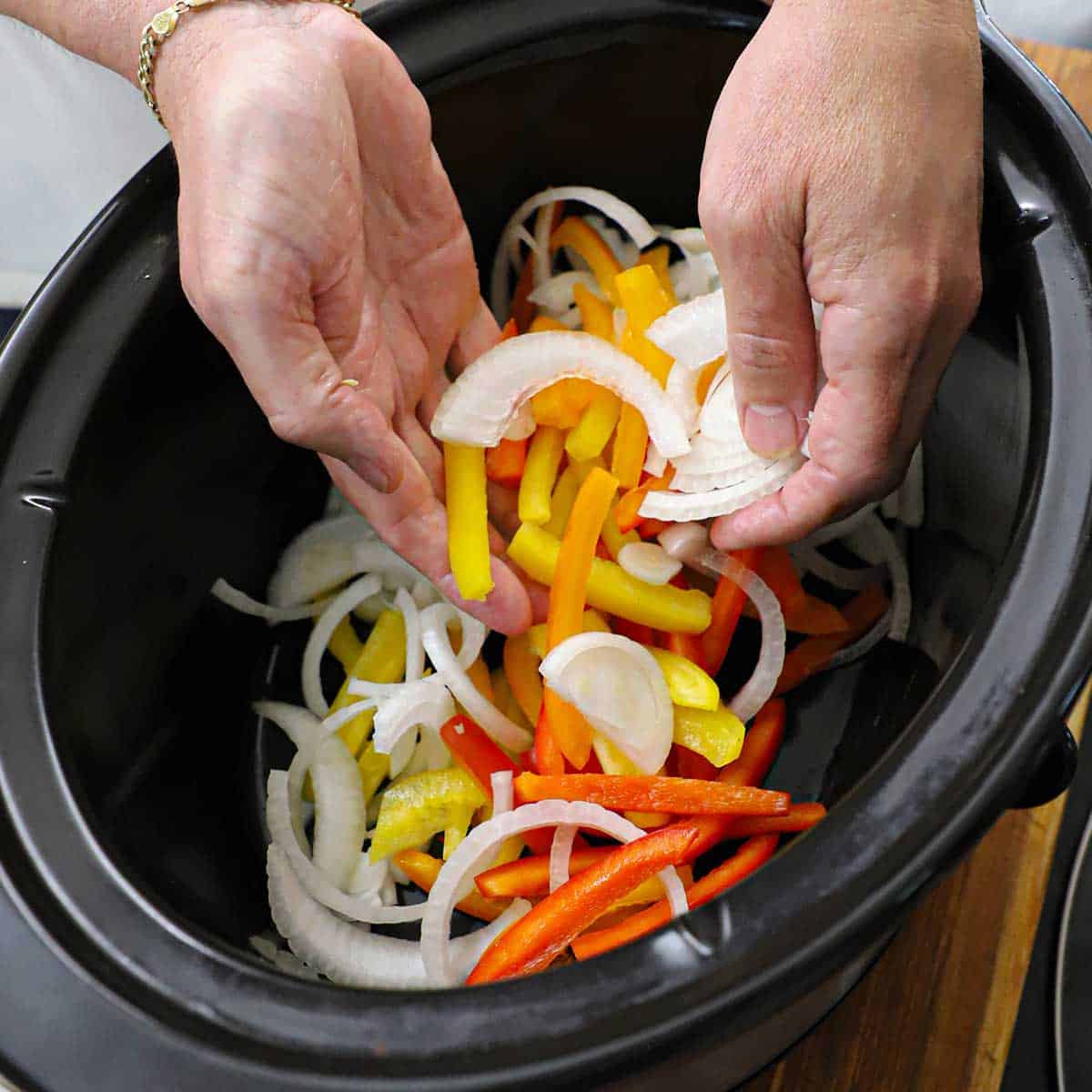 A person using his hands to drop sliced bell peppers and onion into the bottom of a slow cooker.
