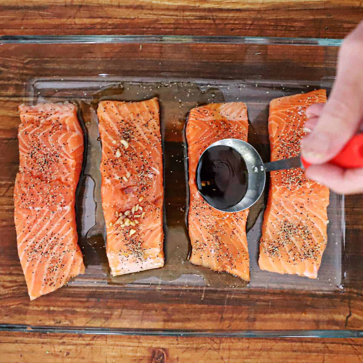 A person using a small ladle to pour teriyaki marinade over four uncooked salmon fillets that have been seasoned with black pepper in a glass baking dish.