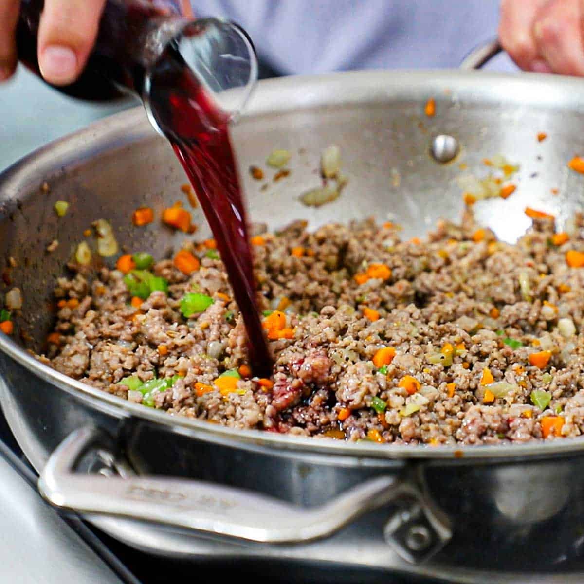 A person pouring red wine from a small glass carafe into a skillet filled with cooked sausage, ground beef, and a mirepoix in a large silver skillet.