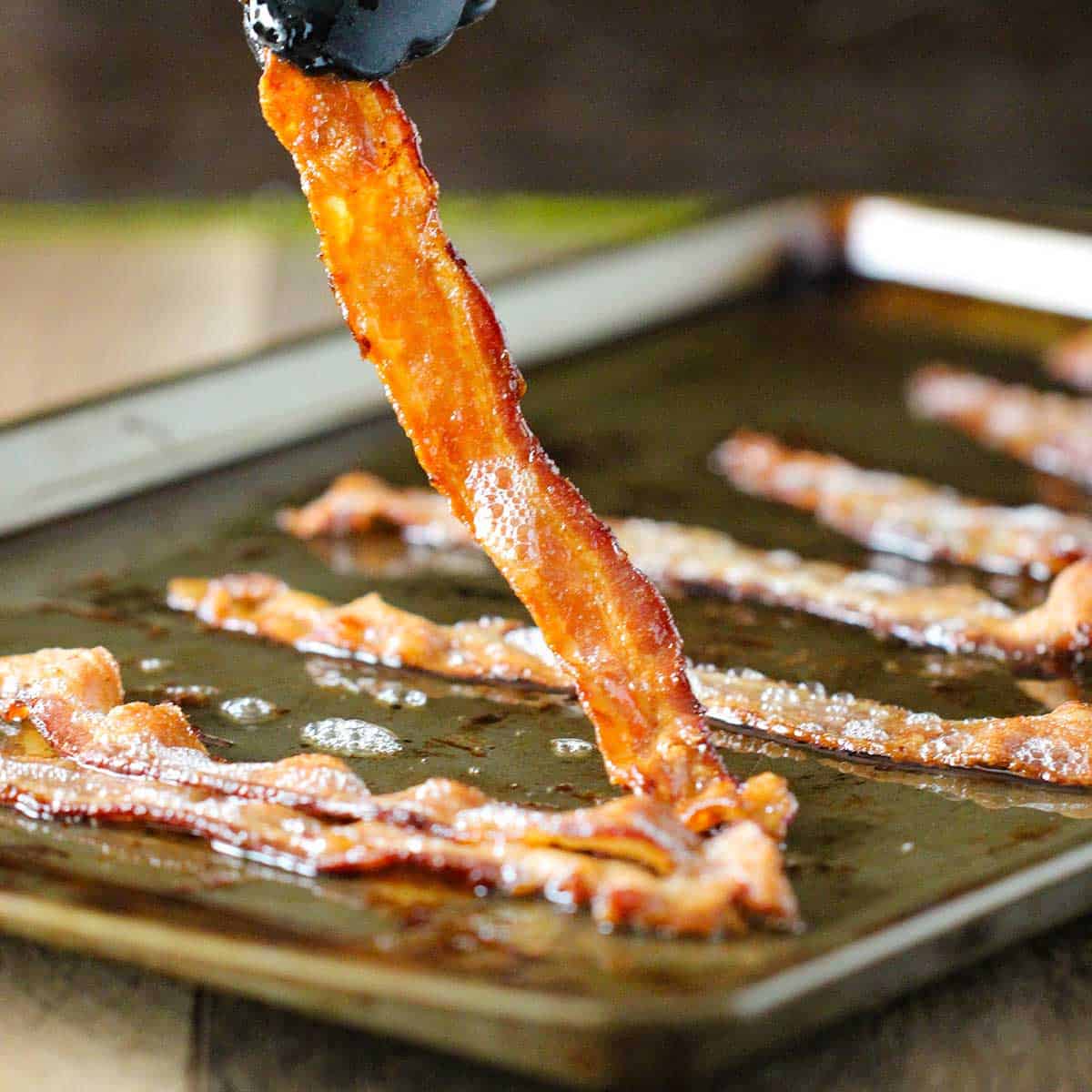 A person using a pair of tongs to lift crispy bacon from a baking sheet that the bacon was baked on.