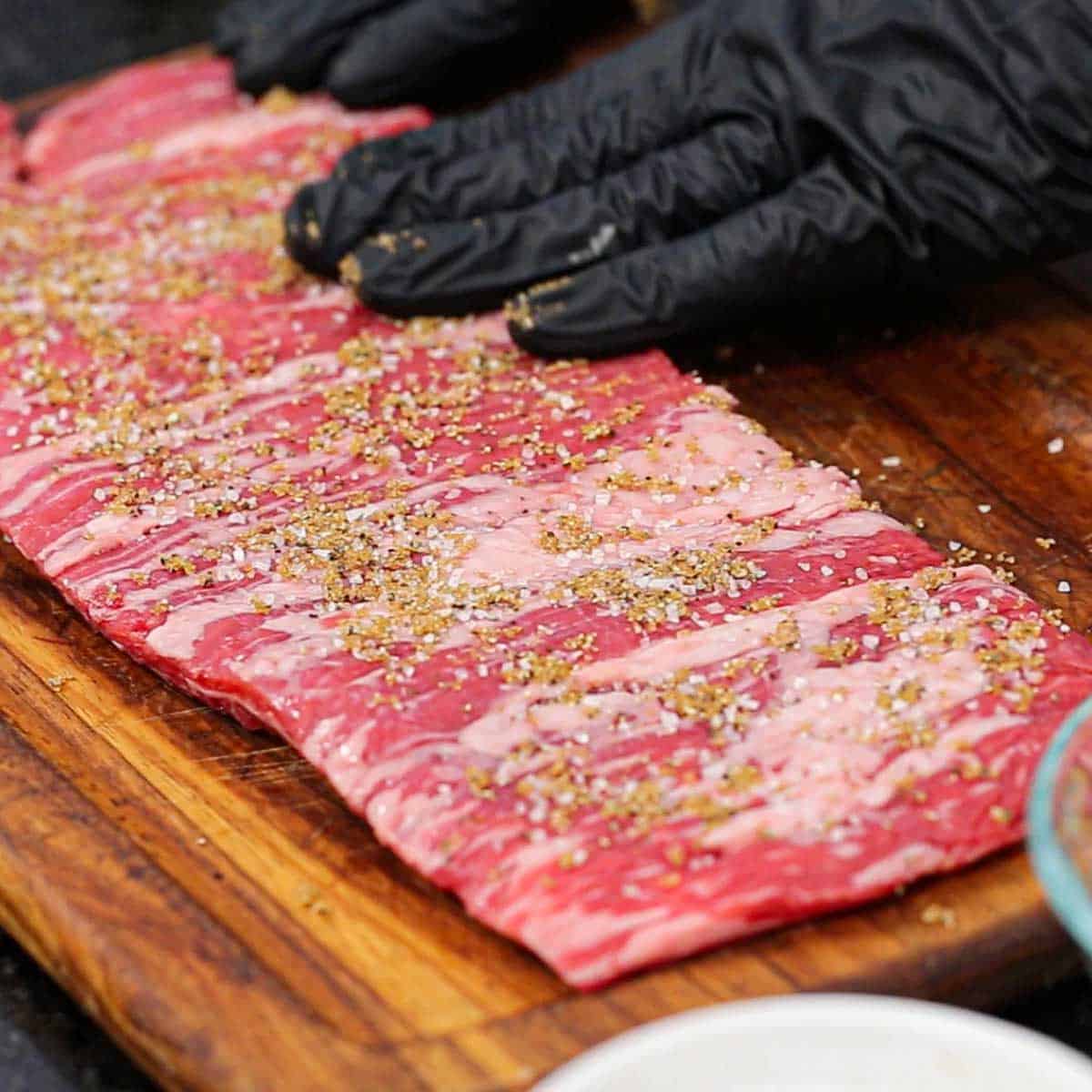 A person wearing black latex gloves pressing a brown sugar and salt seasoning rub onto an uncooked skirt steak on a wooden cutting board.