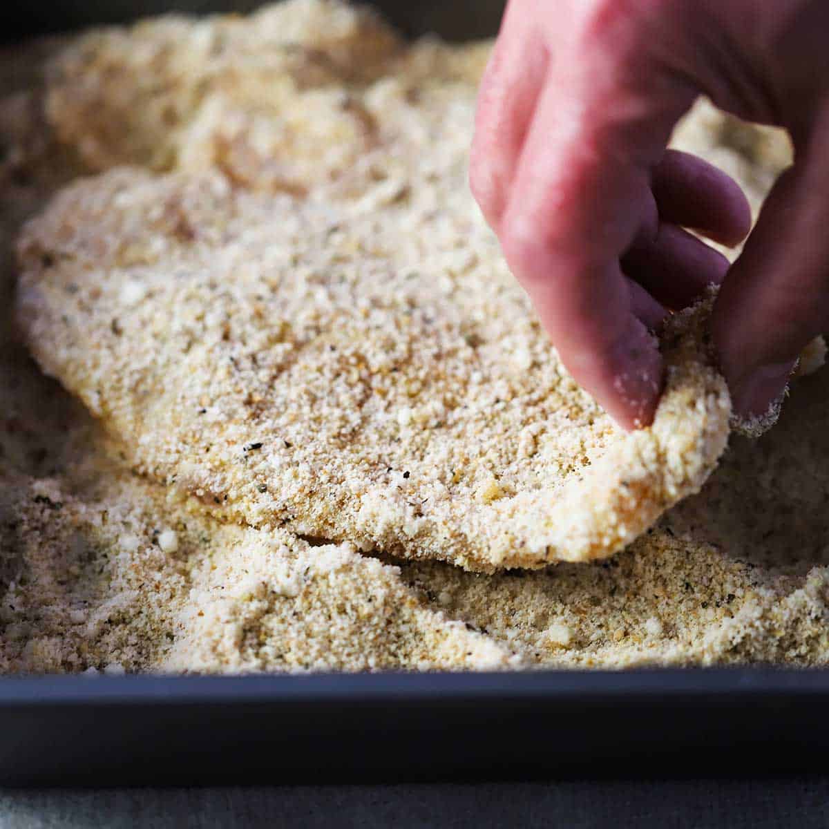 A person dredging an egg coated thin chicken cutlet through a mixture of breadcrumbs and Parmesan cheese in a metal pan.