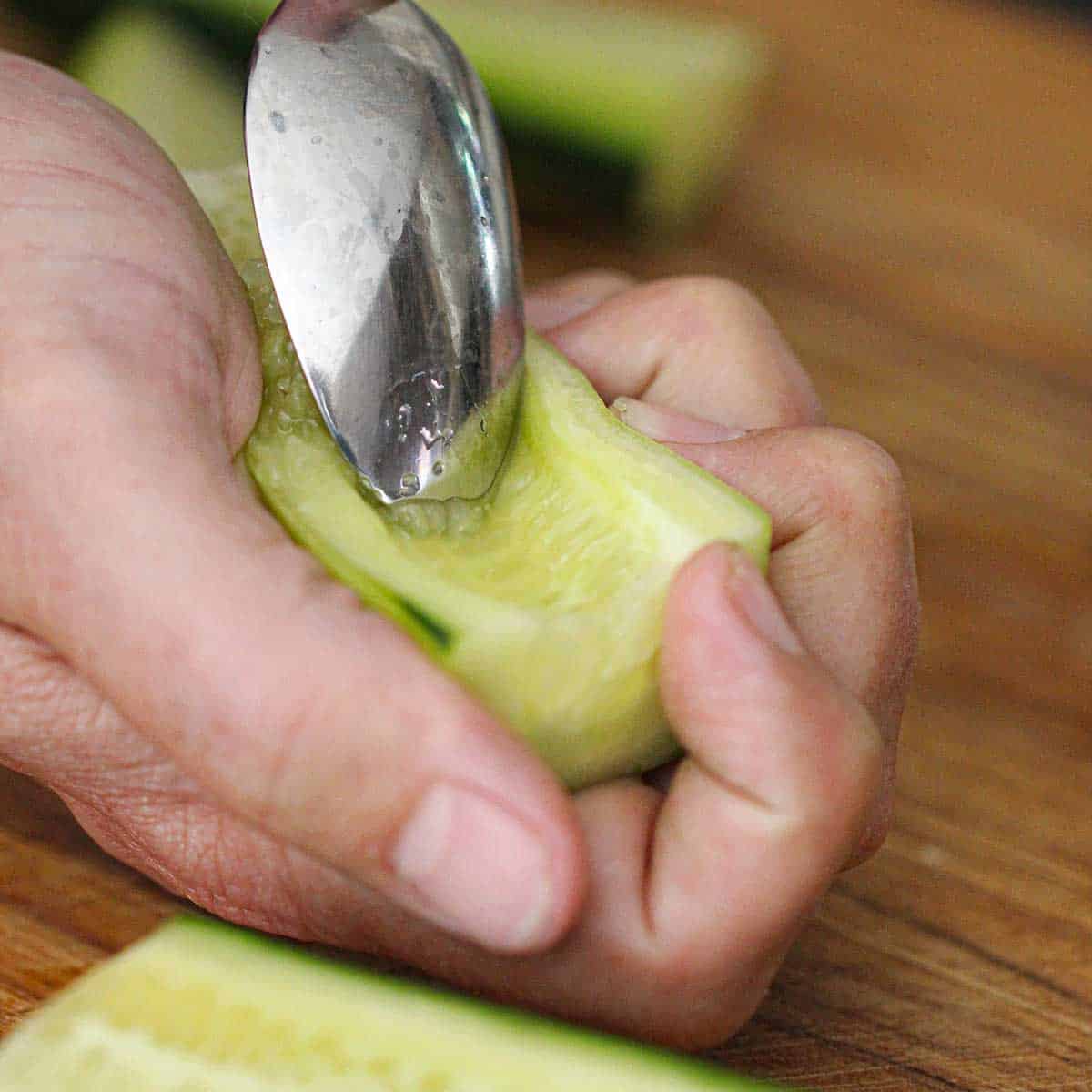 A person holding a halved cucumber in his hand and using a spoon to scoop out the seeds with his other hand.