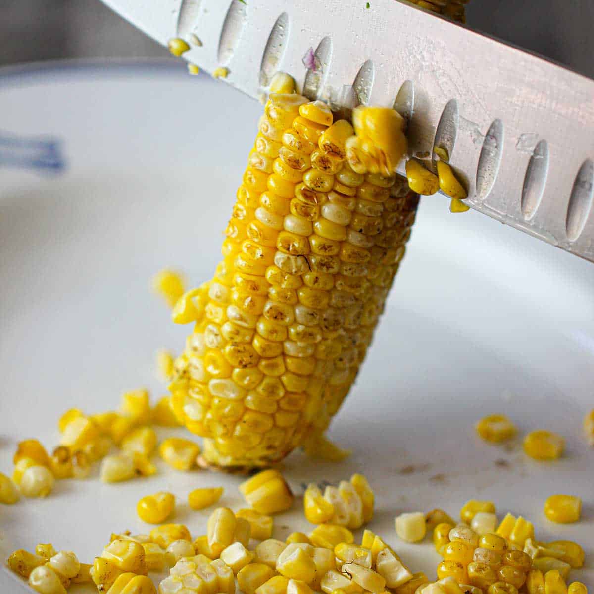A person using a large chef's knife to cut away grilled corn kernels from the cob into a large white platter.