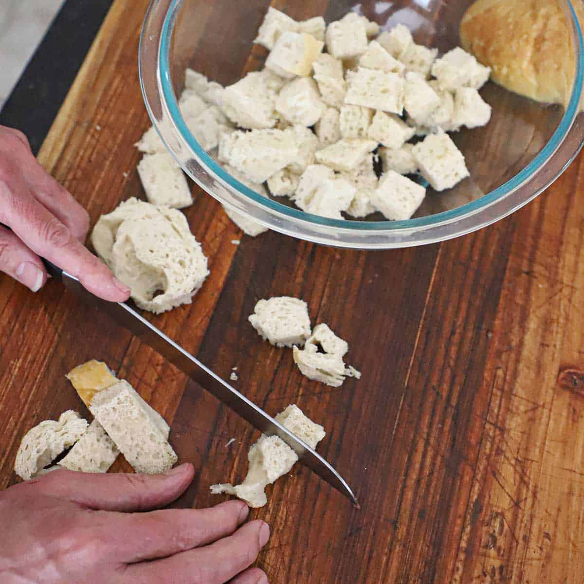 A person using a large serrated knife to cut sliced Italian bread into small cubes with a glass bowl nearby filled with previously cubed bread.
