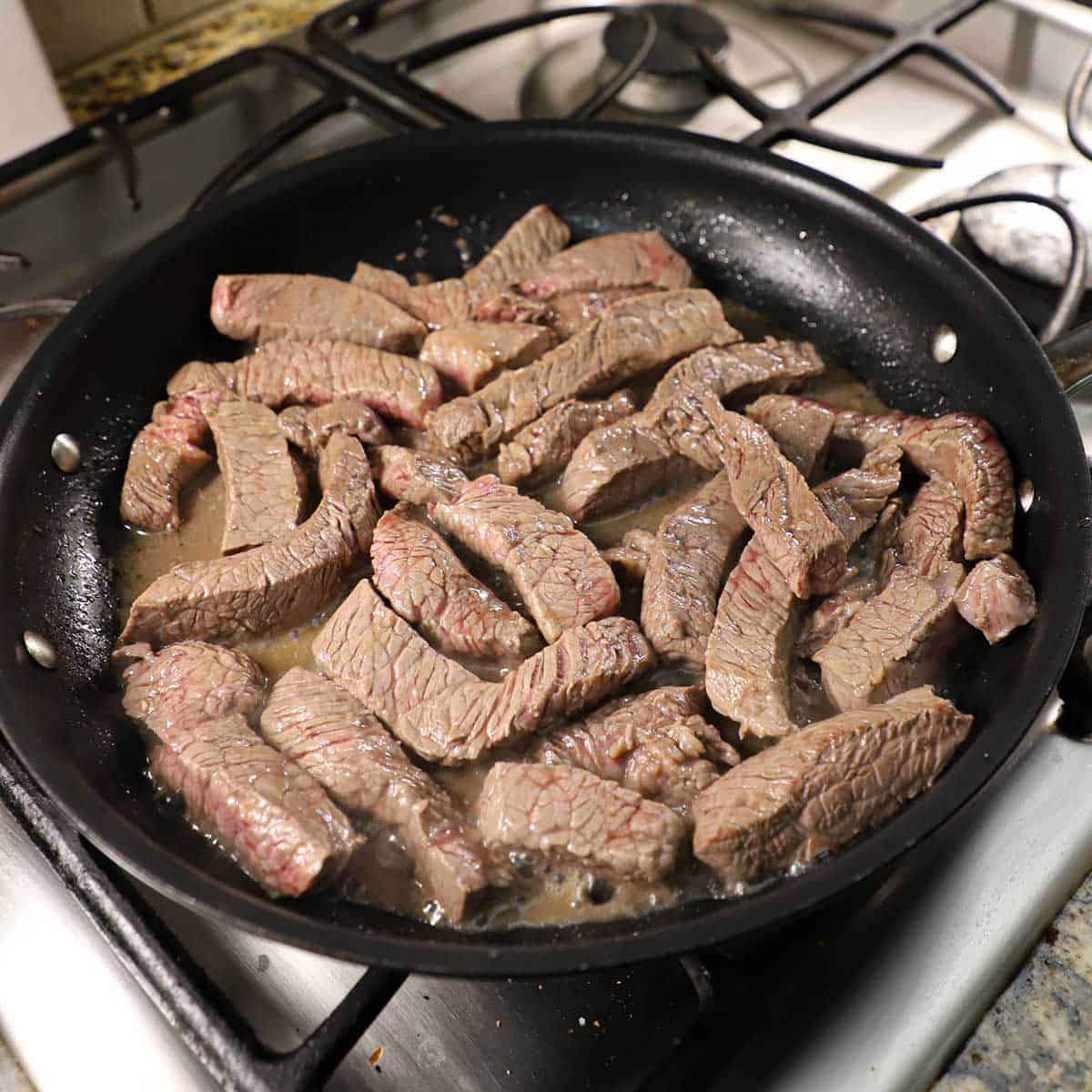 Slices of sirloin simmering in a large black non-stick skillet on a gas stove.