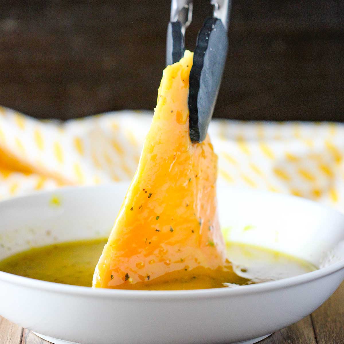 A person using a pair of tongs to lift a thin boneless chicken breast from a shallow white bowl filled honey mustard marinade.