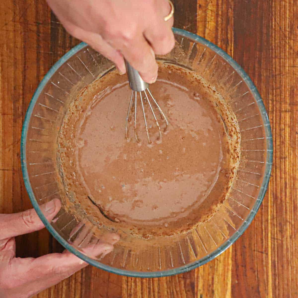 A person whisking a chocolate custard sauce in a glass bowl sitting on a wooden cutting board.
