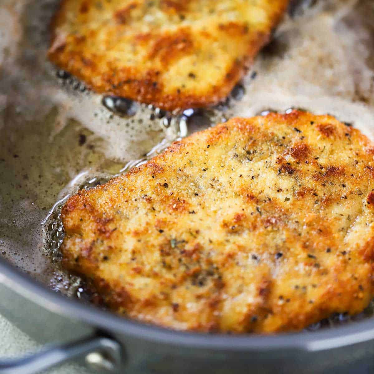 Two thin chicken cutlets that have been coated with breadcrumbs, cheese, and seasonings are being lightly fried in a skillet filled with simmering oil and butter.