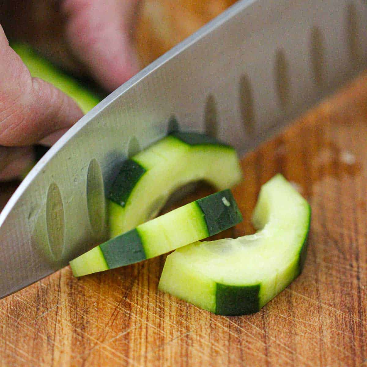 A person using a chef's knife to slice a halved cucumber into slices on a wooden cutting board.