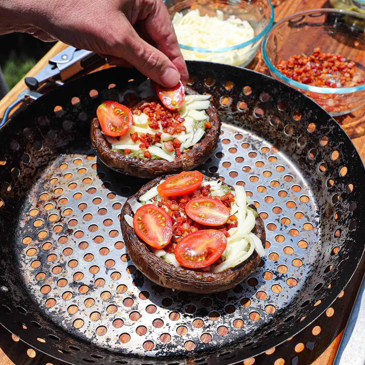 A person building grilled portobello blt pizzas on a metal grill pan consisting of pesto sauce, then shredded cheese, crispy pancetta, and topped with sliced cherry tomatoes.