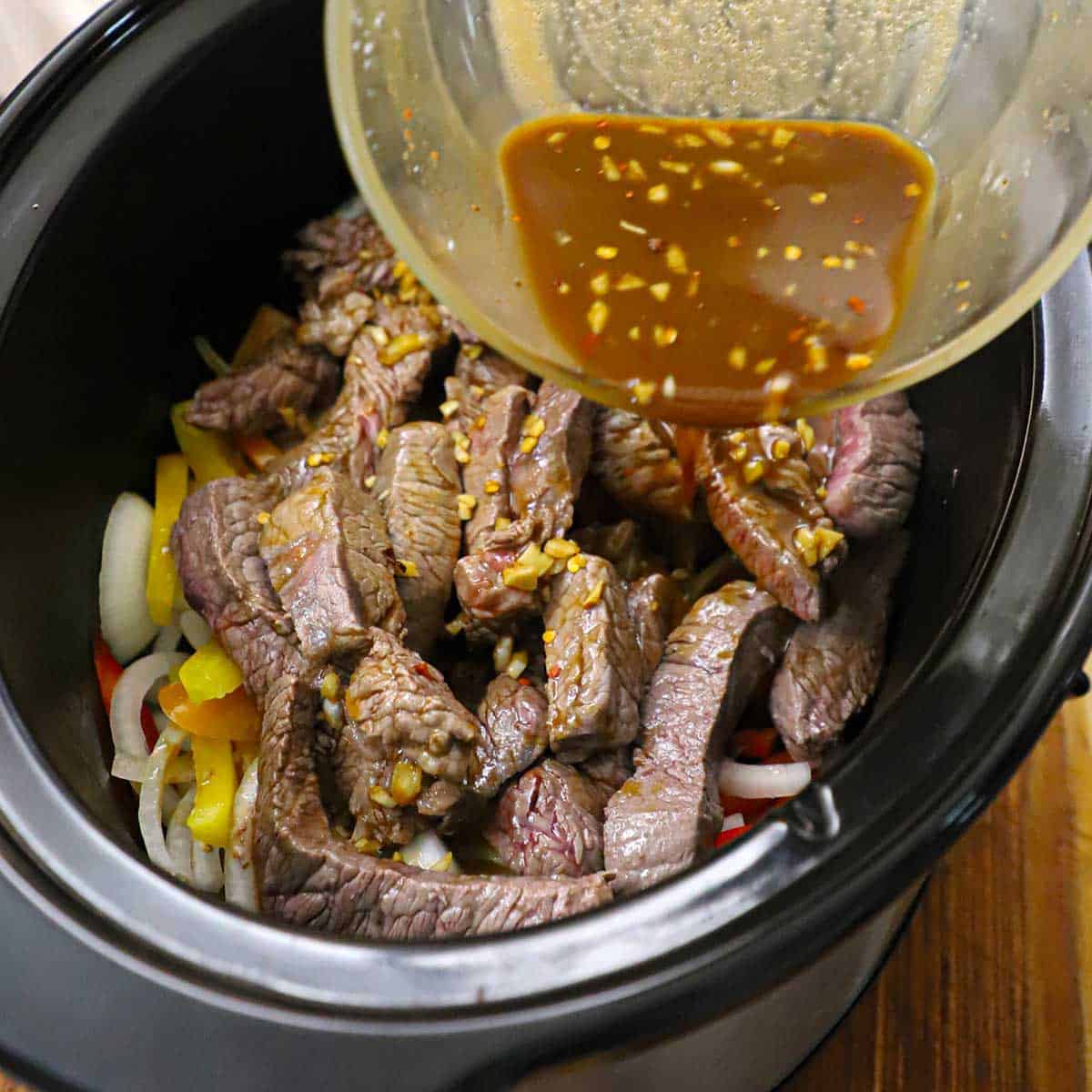 A person pouring a brown pepper steak sauce over seared pieces of sirloin and sliced bell peppers and onion in the bottom of an oval slow cooker.