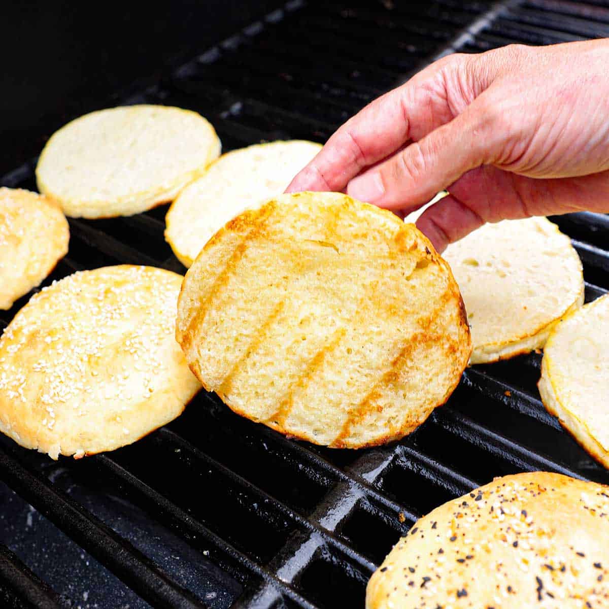 A person holding up a homemade hamburger bun that is being toasted, along with several other buns, on a gas grill.
